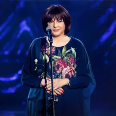 Grażyna Łobaszewska w kwiecistej sukni, gotowa zaśpiewać swój przebój na tle niebieskich świateł.