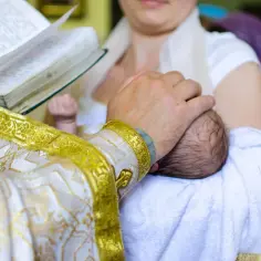 Dłoń duchownego w złotej szacie błogosławi niemowlę. To ważny moment, pierwszy sakrament w życiu dziecka.