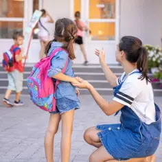 Mama żegna córkę przed szkołą. Czy zerówka jest obowiązkowa? Dzieci z plecakami idą na zajęcia.