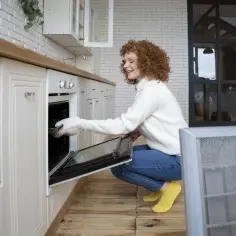 Kobieta w żółtych skarpetkach i białym swetrze, z rudymi lokami, klęczy przy otwartym piekarniku. To jej pierwsze użycie piekarnika, z ekscytacją wkłada do niego naczynie.