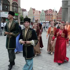 Ludzie w tradycyjnych strojach, przypominających stroje karaimskie, spacerują po brukowanym placu.