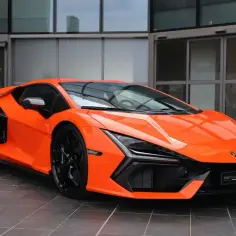 Pomarańczowy Lamborghini Revuelto z czarnymi felgami stoi przed nowoczesnym budynkiem.