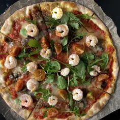 Pyszna pizza z krewetkami, pomidorkami i rukolą, podana na papierze do pieczenia.