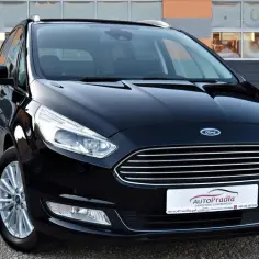 Czarny Ford Galaxy MK3 z chromowanymi elementami, zaparkowany na żwirowym podjeździe przed budynkiem z pomarańczowo-szarymi pasami.