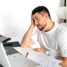 Zamyślony mężczyzna przy biurku z laptopami i dokumentami, zastanawia się, czy umowa zlecenie wlicza się do stażu pracy.