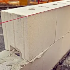 Białe pustaki z widocznymi otworami, ułożone na zaprawie. Czerwona nić murarska wyznacza linię budowy, pomagając sprawdzić wymiary pustaka białego.