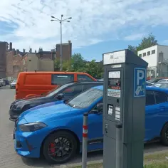 Parking przy Teatrze Rozrywki w Chorzowie. Błękitny samochód i parkometr na tle budynków i pomarańczowego vana.