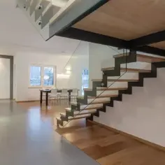 Nowoczesne schody wewnętrzne z drewnianymi stopniami i szklaną balustradą, prowadzące do jasnego wnętrza z kuchnią i jadalnią.