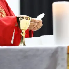 Kapłan w czerwonej szacie, trzymający hostię, przygotowuje się do komunii. Obok kielichy i świeca. Dyskusja o podatek na kościół trwa.