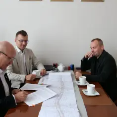 Trzech mężczyzn przy stole omawia mapę i dokumenty. Jeden z nich podpisuje umowę międzynarodową.