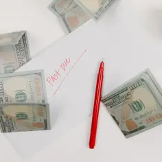 Na stole leżą banknoty 100 USD, czerwony długopis i notatnik. Na kartce papieru widnieje napis "Past Due". To może być faktura imienna z zaległą płatnością.
