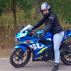 Kobieta w kasku i kurtce na niebieskim motocyklu Suzuki 125 szosowo-turystycznym, gotowa na przejażdżkę po leśnej drodze.