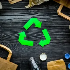 Zielony symbol recyklingu otoczony przez papierowe torby, plastik i butelkę na czarnym tle.