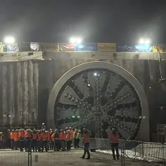 Maszyna TBM gotowa do drążenia najdłuższego tunelu w Polsce. Grupa budowlańców w pomarańczowych kamizelkach obserwuje potężne wiertło.