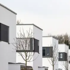 Nowoczesny apartamentowiec z białymi fasadami i szarymi oknami, otoczony nagimi drzewami.