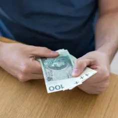 Dłonie liczące polskie banknoty 100 zł na drewnianym stole.