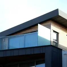 Nowoczesny budynek z drewnianą elewacją i szklanym balkonem. Okap dachu wystaje na tyle, by chronić przed deszczem.
