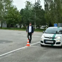 Mężczyzna ustawia pachołek na placu manewrowym przed egzaminem praktycznym kat A. Biały Fiat Panda z zielonym pasem i literą L na dachu czeka na ruch.