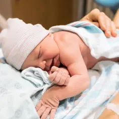 Śpiący noworodek w szpitalnym łóżeczku, otulony kocykiem. Widać jego małe rączki i główkę w czapce. To pierwsze chwile po narodzinach, jeszcze przed pierwszym wypróżnieniem smółki.