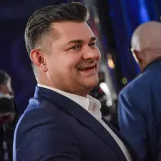 Uśmiechnięty Zenek Martyniuk w garniturze, być może rozmyśla o swoim majątku. W tle fotograf i inni mężczyźni.