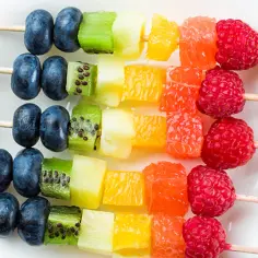 Kolorowe szaszłyki z owocami: borówki, kiwi, ananas, pomarańcza, grejpfrut i maliny ułożone w tęczowym porządku.