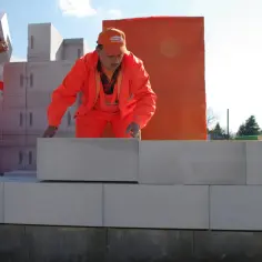Dwóch budowlańców w pomarańczowych strojach układa białe pustaki. Jeden niesie pustak, drugi go kładzie.