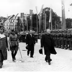 Urzędnicy w mundurach i ksiądz idą w defiladzie, w tle flagi z swastykami. Orawa.