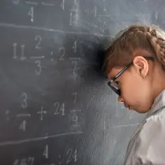 Dziewczynka z okularami opiera czoło o tablicę, zmagając się z zadaniami matematycznymi. Widać jej problemy z nauką i zapamiętywaniem.