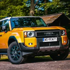 Pomarańczowy Toyota Prado z żółto-czerwonym pasem na boku, zaparkowany na leśnej drodze obok drewnianej chatki.