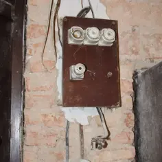 Stary bezpiecznikowy rozdzielacz elektryczny, przygotowany do wymiany instalacji elektrycznej. Ceglana ściana i drewniane drzwi.