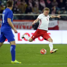 Piłkarz w białej koszulce i czerwonych spodenkach kopie piłkę na boisku.