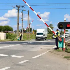 Przejazd kolejowy z zaporami w dół, sygnalizacja czerwona. Stoją pracownicy kolei w kamizelkach. W tle biała ciężarówka i inne pojazdy.