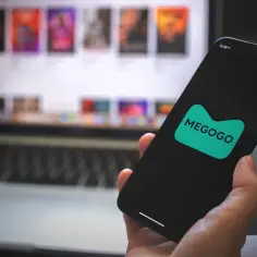 Dłoń trzyma smartfon z logo MEGOGO. Na ekranie laptopa w tle widać okładki filmów.