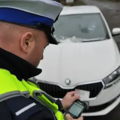 Policjant w kamizelce odblaskowej sprawdza dokumenty kierowcy przy białym samochodzie. Prowadzenie pojazdu pod kontrolą.