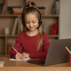 Uśmiechnięta dziewczynka w słuchawkach z mikrofonem pisze w zeszycie. Obok laptop i kubek. Zastanawia się, nauczanie indywidualne ile godzin jest potrzebne.