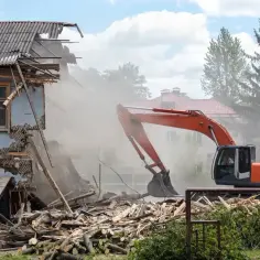 Pomarańczowy koparka wykonuje demontaż zniszczonego budynku, unosząc tumany kurzu.