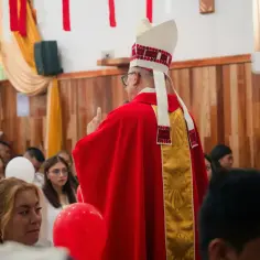 Biskup w czerwonej szacie i ozdobnym nakryciu głowy błogosławi zgromadzonych wiernych.
