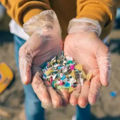 Ręce w rękawiczkach trzymają garść kolorowych drobinek plastiku, przypominających mikroplastik w kosmetykach, znalezionych na plaży.