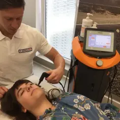 Fizjoterapeuta wykonuje zabieg indiba na twarzy pacjentki. Urządzenie z ekranem monitoruje parametry terapii.
