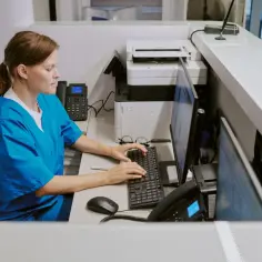 Pielęgniarka w niebieskim uniformie pracuje przy komputerze. Najnowsze wiadomości o podwyżkach dla pielęgniarek mogą być powiązane z jej pracą.