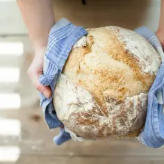 Ręce trzymają świeżo upieczony, złocisty bochenek chleba owinięty w niebieski ręcznik.