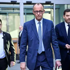 Friedrich Merz, prezydent Niemiec, w niebieskim garniturze, wychodzi z budynku, otoczony ochroniarzami.
