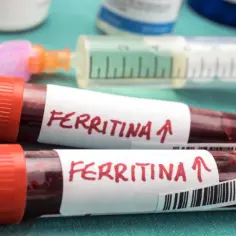 Dwie probówki z krwią oznaczone "Ferritina" i strzałką w górę, sugerujące potrzebę podniesienia ferrytyny. W tle strzykawka i fiolki.