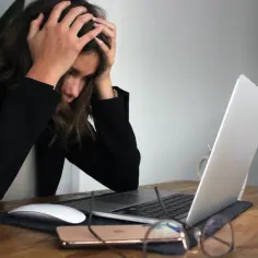 Kobieta z rękami na głowie przy laptopie, telefonie i okularach. Widać zmęczenie i myśl o porzuceniu pracy.