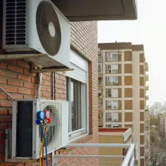 Dwa urządzenia klimatyzacyjne zamontowane na ceglanej ścianie bloku mieszkalnego, z widokiem na inne budynki.