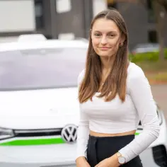 Uśmiechnięta nastolatka z długimi włosami stoi przed białym samochodem. Niedługo skończy 17 lat i marzy o swoim pierwszym **prawie jazdy**.