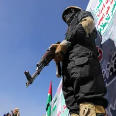 Mężczyzna w czarnym stroju i masce trzyma karabin. W tle transparent z arabskim napisem, symbolizujący ruch Huti.