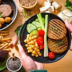 Talerz z wegańskimi burgerami, ciecierzycą, pomidorkami i ogórkiem. W tle inne wegańskie produkty.