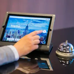 Dłoń dotyka ekranu tabletu z widokiem na Nowy Jork, gdzie można sprawdzić, ile trwa doba hotelowa. Obok stoi dzwonek hotelowy.