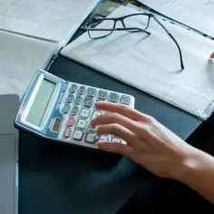 Dłoń wpisująca dane na kalkulatorze, obok dokumenty i okulary.